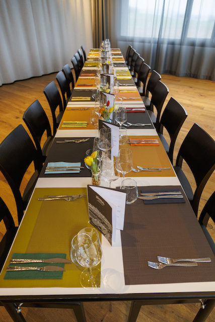 Langer gedeckter Tisch mit Stühlen, Gläsern, Besteck und Menükarten für eine Feier. Die Tischsets sind in verschiedenen Farben gehalten. Frische Tulpen in kleinen Vasen als Dekoration. Das Licht ist sanft und natürlich.